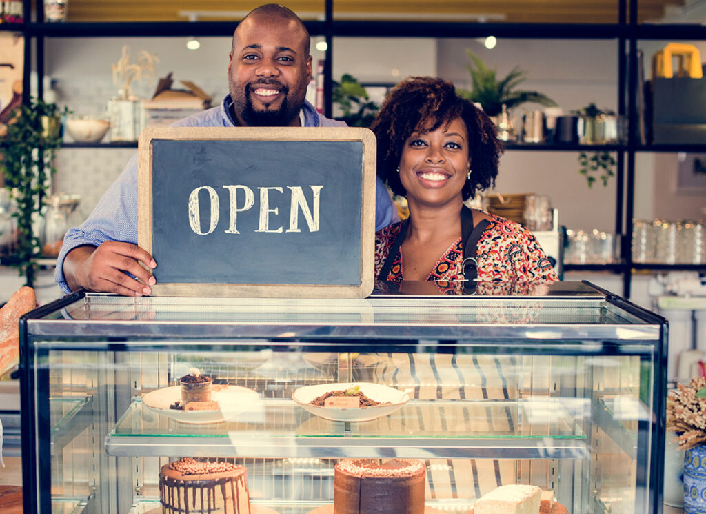 Business Tax Solutions - Happy Couple Stand Together in Their Bakery Holding an Open Sign